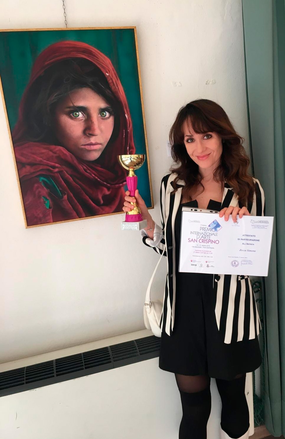 Simona Zecca regge una coppa vinta per il dipinto che ritrae la famosa ragazza afgana, appeso al muro affianco a lei 